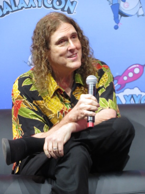 Weird Al sitting onstage with mic, smiling and listening.