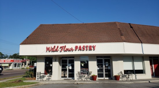WildFlour Pastry restaurant in a strip mall, red logo on white wall, brown shingled roof. Three empty tables outside.