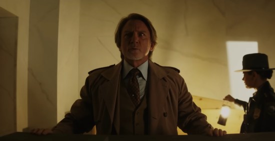 A confused Benoit Blanc stands over a casket, resting his hands on the lid. At right is State Trooper Mila Kunis.