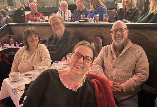 Four people seated at a dinner theater table, smiling at camera.