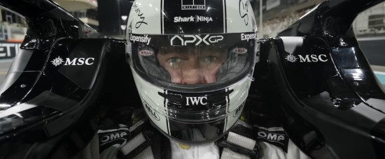 Brad Pitt as a race car driver in black-and-white suit and helmet, sitting in black car covered in logo stickers.