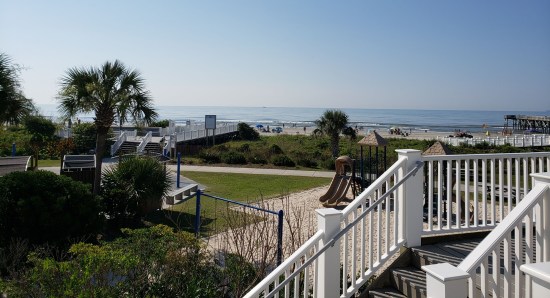 distant ocean and beach past a deck staircase in the foreground and a playground not far off.