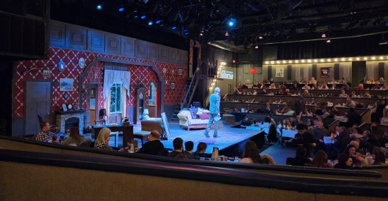 Nicely dressed gentleman addresses a theater from a stage containing 1940s furniture and inn decor.