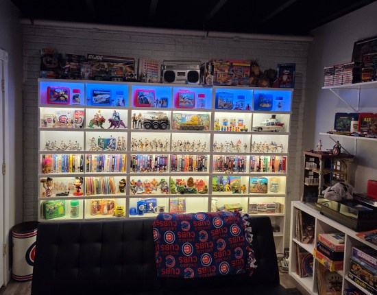 White shelves filled with toys and movies, and a couch with a Chicago Cubs blanket on it.
