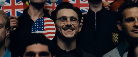Timothee Chalamet in period-piece mustache holding ping-pong paddle with American flag design, standing amid other players with paddles bearing their own homeland's flags.