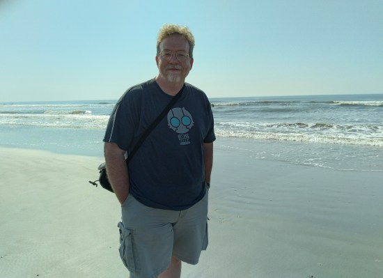 me on a white beach by the ocean, wearing off-white shorts and an Atomic Robo T-shirt.
