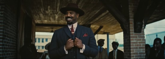Michael B Jordan in red hat and Sinners suit, grinning and surveying his hometown.