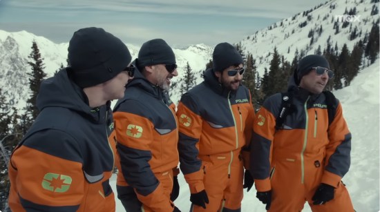 Four billionaires in black and orange snowsuits and black winter caps, goofing on a snowy mountainside.