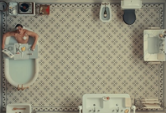 Very large 1920s bathroom with nothing in the middle except white-print floor. At upper left, Benicio Del Toro is in a bathtub, looking up.
