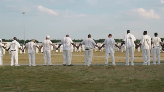 Inmates dressed in white join hands in a circle in a grassy prison courtyard.