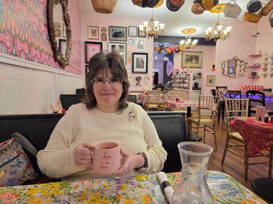 Anne sitting in a very pink restaurant, holding a mug that says "Love is love". Baskets hang from the ceiling.