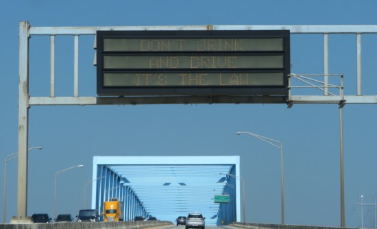 Interstate bright with rectangular blue-girdered bridge. Overhead signboard lights up to say, "Don't drink and drive, it's the law."