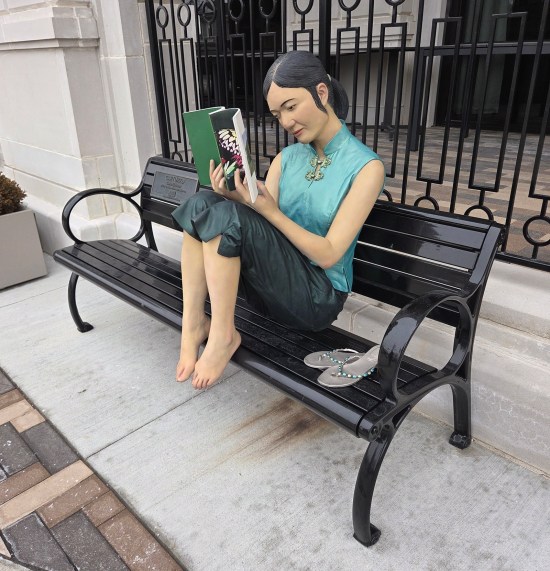 Statue of Asian woman curled up on park bench, reading a softcover nature book.