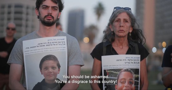 Israeli man and woman stand silently and mournfully, holding posters with pics and bios of dead Palestinian children.