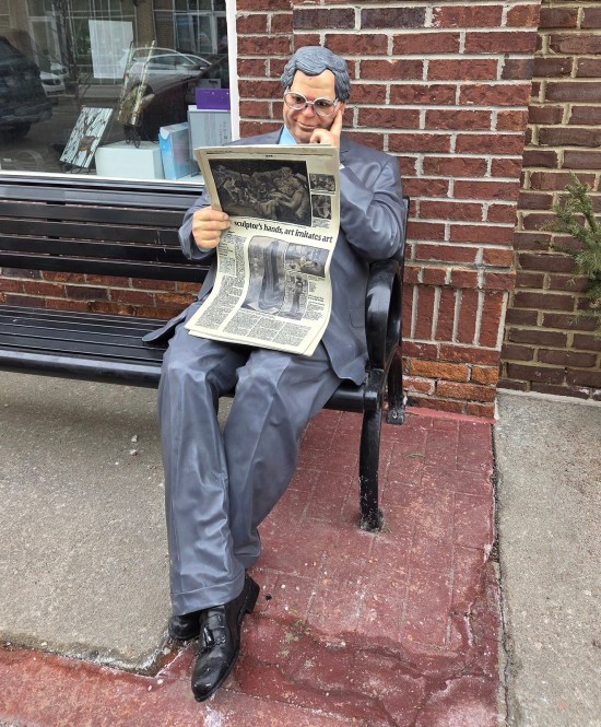 Statue of bespectacled businessman reading newspaper on bench. The arts section is turned toward us with an article about a sculptor.