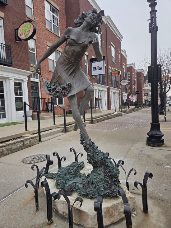 Statue of woman in negligee sniffing one rose, holding a bouquet in her other hand, flying in the air and leaving a trail of roses behind her.