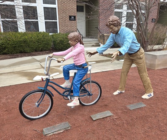 Statue of tiny girl in pink turtleneck sweater and blue jeans nervously riding a girl's bike with banana seat. Dad walks behind her, arms outstretched in case she collapses.