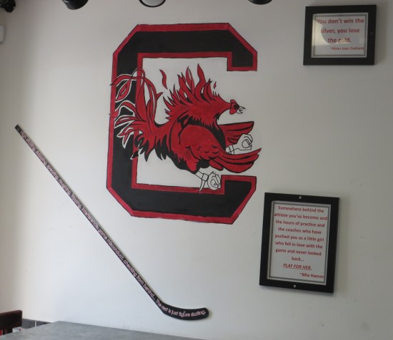 Black and red C with a red chicken inside it, painted on a restaurant wall. Also hung by it are a hockey stick and framed inspirational quotes.