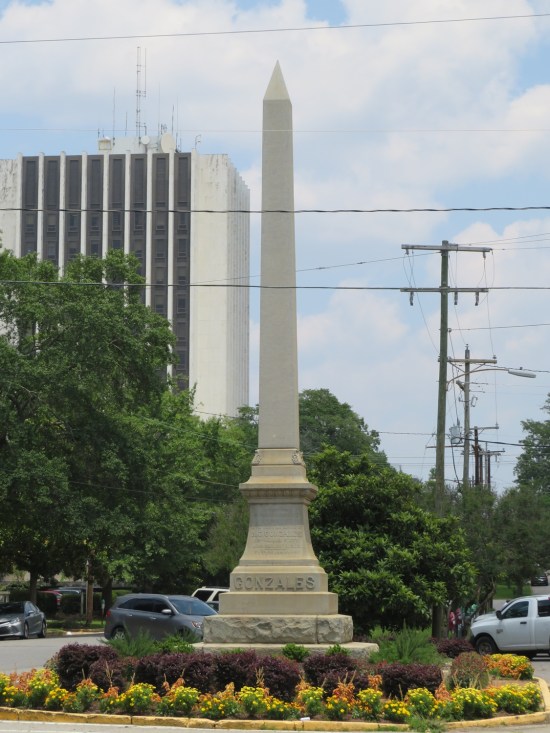 Stone obelisk in the center of a street that says "GONZALES" on one side. Flowers bloom around the base.