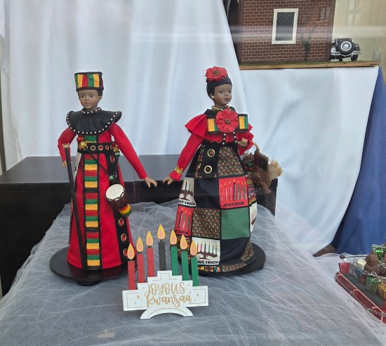 Pair of Kwanzaa dolls in red and green standing in front window on white table.