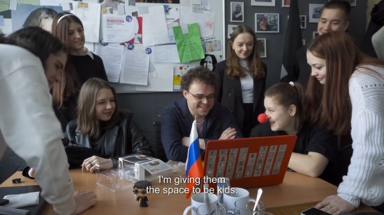 Happy Russian teacher at desk surrounded by happy teenage students. Subtitle reads, "I'm giving them the space to be kids."