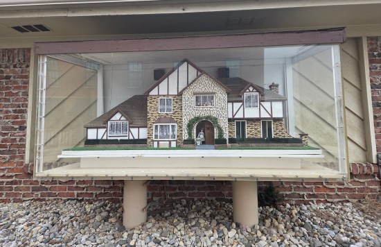 Large brown and white miniature house in a glass display on two poles outside the museum.
