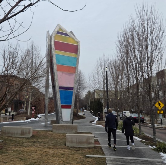 sculpture of three disproportionately tall pentagons with colored stripes, installed along a bricked trail with two passing walkers.
