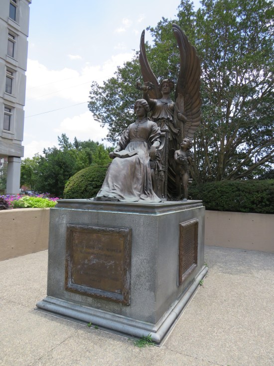 Statue of 19th-century woman sitting with book. Behind her is an angel holding a laurel crown over her head, and a cherub.
