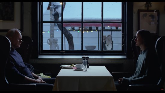 Stellan Skarsgard and Renate Reinsve share a darkened restaurant table in front of a window at sundown, He has an empty plate, her side is empty. Both have neutral expressions.