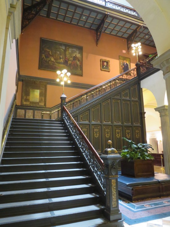 Staircase up to second floor of statehouse. Orange walls, some paintings.