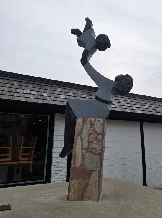 Stylized statue of Black mother happily swinging her toddler high in the air by both arms. Both are faceless. A restaurant is behind them. 