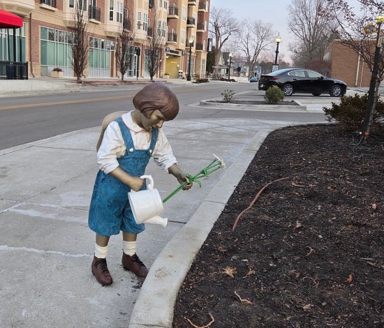 Statue of Buster Brown in blue overalls with a watering can and three plucked long-stem flowers that can't just replanted that easily.