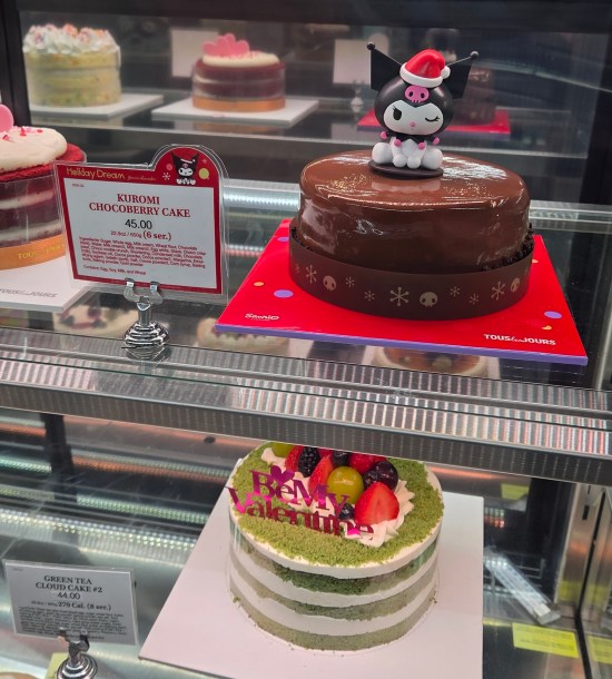 Display case with two cakes: a green and white one with fruit on top and the words "Be My Valentine", and one chocolate cake topped with a Kuromi figurine on top, which I had to Google.