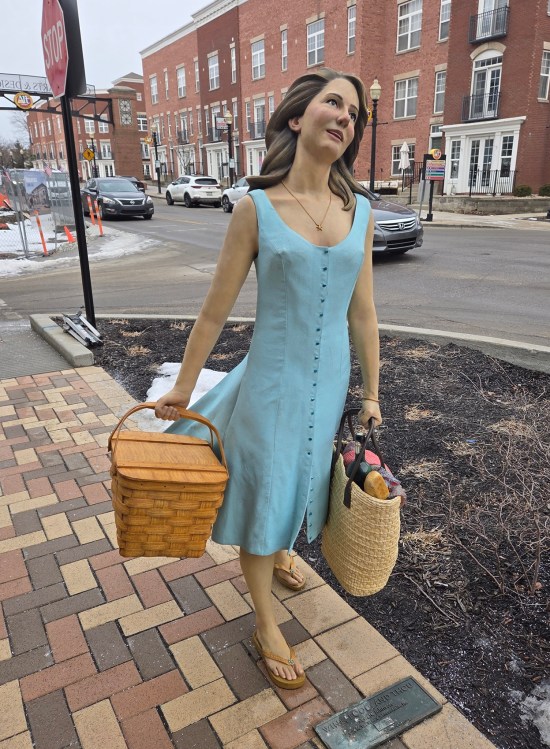 Statue of Jewel Staite in a blue sundress carrying a picnic basket and a tote bag full of groceries.