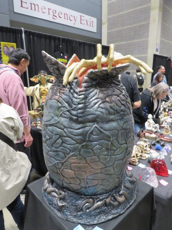 Large replica of hatching egg from the movie "Alien", sitting on a comic-con table in front of a hugely labeled emergency exit.