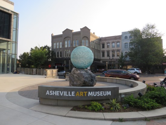 Outdoor pavilion with Asheville Art Museum sign on a low, curved wall. In the middle is a sculpture of a light blue, ridged sphere atop a large, uneven rock.