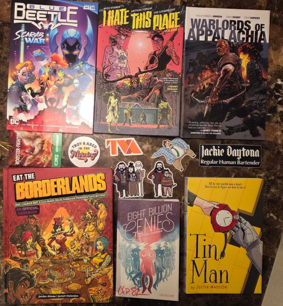 Six graphic novels and comics, plus six stickers, arranged on our dining table.