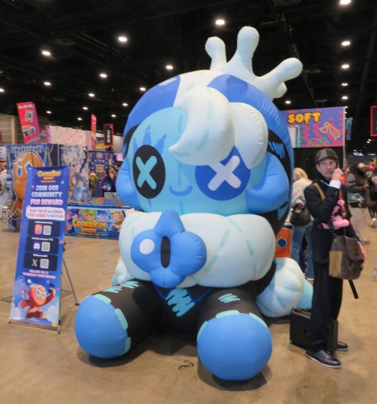 Giant blue inflatable kiddie anime figure with X's on its eyes, sitting in a con booth.