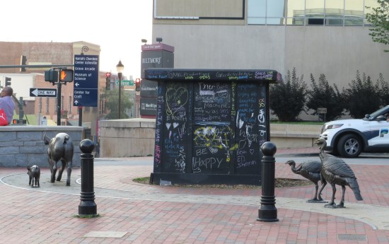 Empty black pedestal on a brick city sidewalk, surrounded by statues of two pigs and two flightless birds.