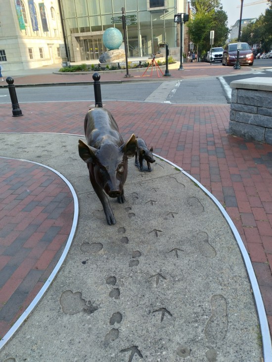 Sculptures of pig and piglet walking on fake wet cement with multiple animal tracks in it.