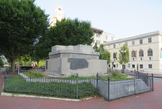 empty white pedestal surrounded by boards, near the end of a small city park.