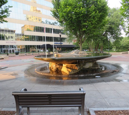 Large, flat-topped bowl-shaped fountain held up by rocks that are lit up by the sunset. Background is an office building with a sign reading "Biltmore".