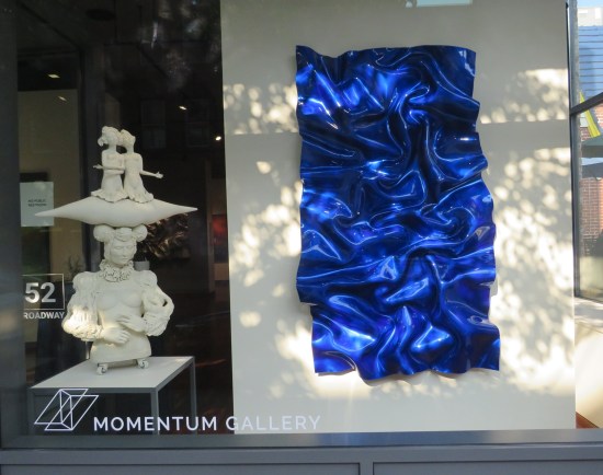 Art gallery front window with two pieces: one all-white Asian sculpture, and one large crinkled sheet of blue foil hanging on a white backdrop.