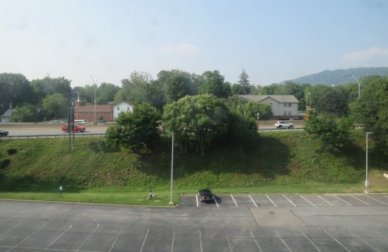 Nearly empty parking lot behind a hotel, A grassy hill and a few trees separate it from an interstate. One grassy mountain is on the horizon.