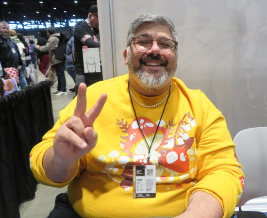 Jarrett Melendez at his C2E2 table, smiling and flashing a peace sign.