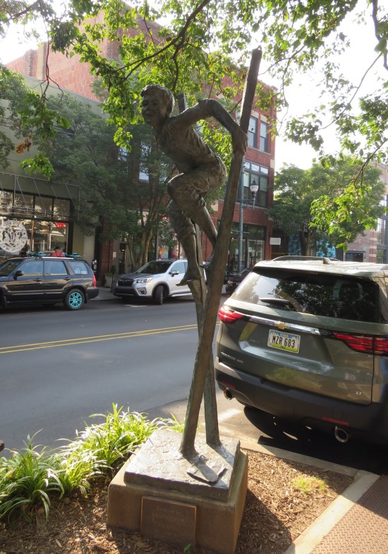 Statue of boy in overalls on stilts, installed near a city sidewalk.