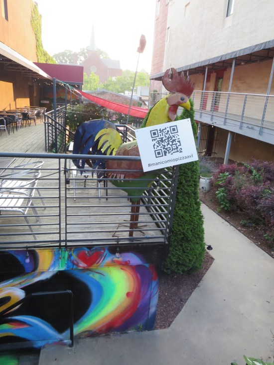 Rooster sculpture perched on a pizza place patio, holding a QR code.