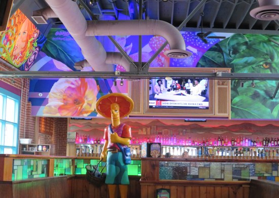 Restaurant interior with pink-lit bar, yellow mushroom man mascot statue, green and purple painted walls, and industrial-chic ceiling pipes.