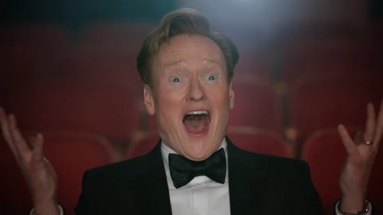 Conan O'Brien sitting in am empty theater, marveling at the screen and holding his hands up in surprise.