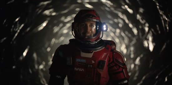 Ryan Gosling in red astronaut armor with a NASA logo, smiling and standing in a dark foil-lined tunnel.